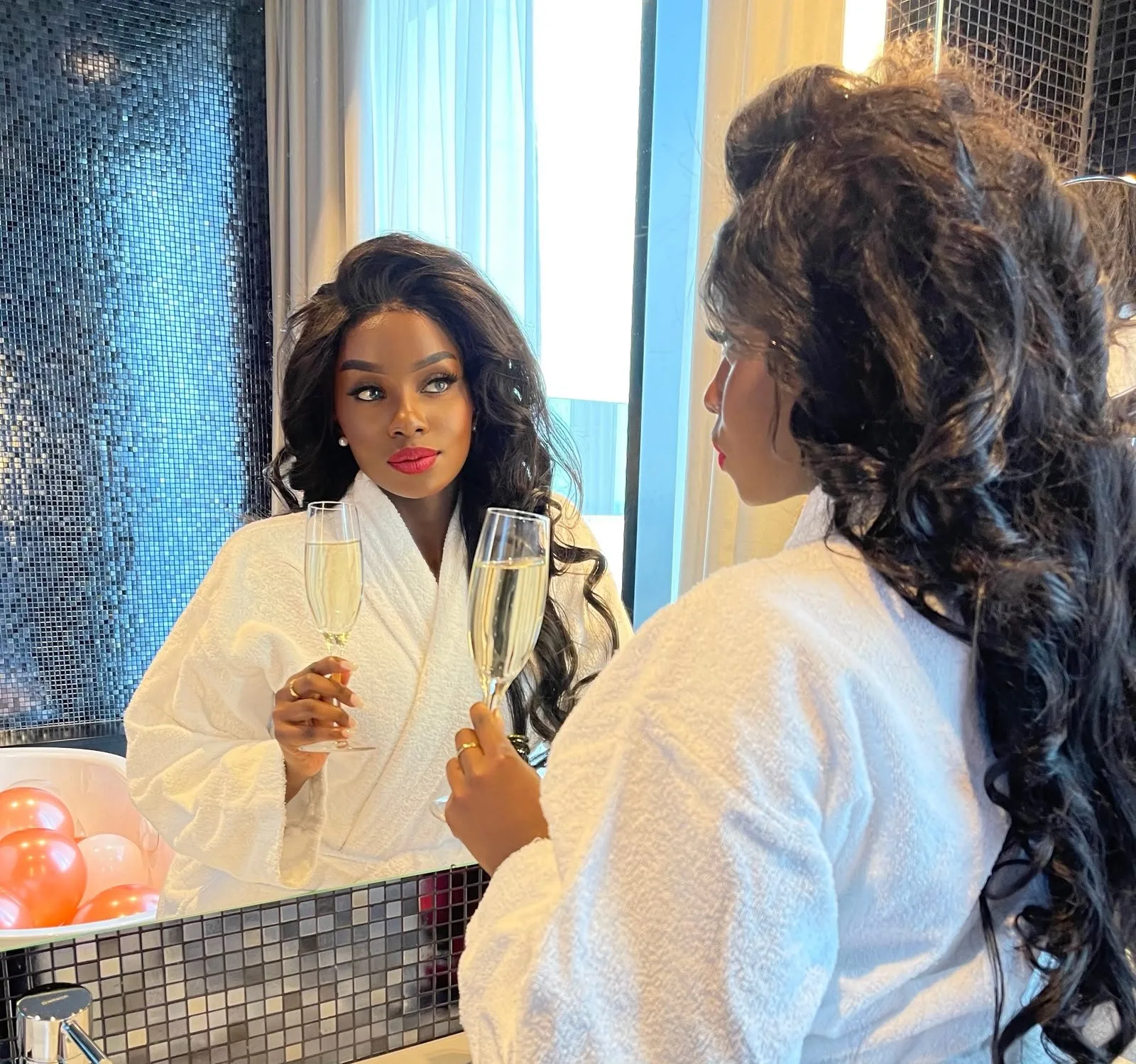 June in a white robe raising a glass of champagne to her reflection